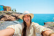 © Daniel - Young woman wearing a hat and striped shirt smiling, holding a phone for a selfie during a summer vacation by the ocean coastline with colorful buildings in the background