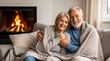 © Robson - Smiling senior couple sitting on sofa wrapped in blanket by fireplace in cozy living room