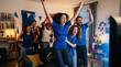 © Ada - Enthusiastic multi-ethnic group of friends wearing blue sports jerseys celebrating a goal while watching a soccer match on TV in a decorated living room at home in the evening.