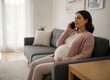© abimas - Pregnant woman sitting on a gray sofa at home talking on her smartphone
