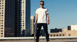 © Lubov - Young man stands on rooftop holding skateboard. He sports buzzed hair sunglasses casual attire. Tall buildings form urban backdrop under clear blue sky. Cool skater enjoys city view relaxed attitude.