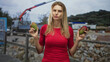 © Krakenimages.com - Woman holding two mangoes, standing on street by a crane and truck near a rocky seawall; calm contemplation.
