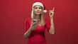 © Krakenimages.com - Blonde woman holding smartphone and pointing finger upward in studio wearing santa hat and red shirt; festive cheer.