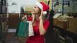 © Krakenimages.com - Woman in santa hat holding green and white shopping bags, smiling and cups ear while looking up in bookstore; holiday joy.