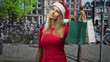 © Krakenimages.com - Woman holding shopping bags by hand, tilting head, wearing santa hat and red top in cobbled street with bicycles and canal; festive shopping.