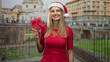 © Krakenimages.com - Blonde woman in santa hat smiles and holds a red gift box in her hand on a city street by historic buildings; festive joy.