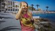 © Krakenimages.com - Woman holding cocktail mug makes ok sign to eye in front of building on sunny seaside promenade; joyful summer fun.