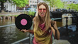 © Krakenimages.com - Blonde woman holds vinyl record with pink label, pumps fist and sticks tongue out while wearing pink sunglasses on street canal bridge; joy celebration.