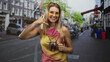 © Krakenimages.com - Woman holding copper cocktail mug and smiling, right hand making call gesture while standing on a busy street lined with parked bikes and buildings; cheerful invitation.