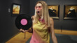 © Krakenimages.com - Blonde woman holding vinyl record with pink label, mouth open, wearing pink sunglasses and yellow dress inside a building gallery with framed paintings; surprise nostalgia.