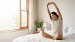 © Xenia - Young woman stretching arms while sitting on bed in the morning. Happy female waking up in bright bedroom. Healthy lifestyle and morning routine concept
