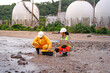 © ultramansk - Two engineers in safety gear collect water samples and input data on a laptop near coastal chemical storage tanks. Field research and pollution monitoring in action.