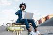 © Westend61 - Spain, Barcelona, young businessman sitting outdoors in the city using cell phone and laptop