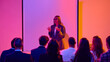 © Lustre Art Group  - Female presenter speaking to business audience with hands raised in neon classroom. Concept of leadership training, workplace learning, soft skills workshop, mentoring and team communication.