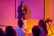 © Lustre Art Group  - Smiling female presenter leading audience discussion in modern neon workshop. Concept of interactive training, business education, team learning, public speaking practice and friendly coaching.