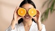© ruangrit19 - Young smiling Asian woman holding fresh orange fruit slices over her eyes for a fun skincare and beauty concept in a bright studio.