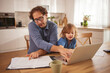 © Stockphotodirectors - A father helps his child with a laptop while surrounded by documents on a table in their cozy kitchen. Sunlight streams through the window, creating a warm atmosphere.
