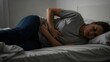© Djemari - A woman lies in bed, curled up and holding her stomach, appearing to be in pain or suffering from abdominal discomfort in a dimly lit room.