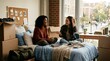 © Pramata - Two diverse female college students laughing and talking while sitting on a bed in a cozy dorm room during move-in day with cardboard boxes.