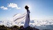 © Catherine - Black woman standing on a rocky mountain peak above the clouds, wearing flowing white fabric lifted by the wind. Bright sunrise light creates an ethereal, peaceful atmosphere with soft golden tones.