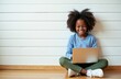 © miss irine - Smiling young black girl learns online using a laptop. Child sits cross-legged on floor typing keyboard, bright smile, focused on education. Technology use for kids learning.