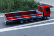 © massimo_g - Camion pianale rosso parcheggiato su strada con area verde e marciapiede sullo sfondo. Ideale per temi di trasporto, logistica, edilizia, consegne e mobilità commerciale.