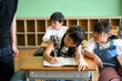 © apiox - 教室で勉強する女子小学生と教え合う友達を傍で見守る男性教師