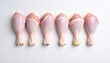 © royaesthetic - Seven raw chicken drumsticks arranged neatly in a horizontal line on white surface, symbolizing culinary preparation and fresh poultry.