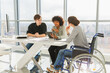 © Юлия Завалишина - Teamwork brainstorm. Diverse multiracial colleagues woman in chair for people with disability working together. Group of people sitting in creative office discussing business. Young professional team