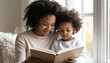 © Neural Canvas - Mother child reading book family parent toddler window home together learning education bonding happiness affection smile reading childhood curly hair casual clothing sofa daylight natural light