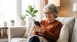 © DhimasPratama - Smiling senior woman with glasses using smartphone on living room sofa