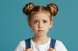 © Pete - Young girl with blonde hair in buns looks confused with worried expression. Child has blue eyes and furrowed brow. She wears blue denim overalls over white t shirt against teal background.