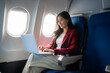 © Thitaporn - Smiling young woman using a laptop while seated by the window on an airplane, showing business travel, remote work, comfort, and modern mobility.