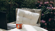 © Anya Kamyshnikova - A wicker chair with a white cushion stands behind a table covered with a light cloth. On the table are a laptop and a red coffee mug, surrounded by blooming pink flowers and green foliage.