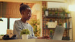 © DC Studio - African American woman works at home during sunset hour, publishing blog article on personal website. Freelancer seeking to build online e-business while refining abilities. Handheld shot.