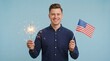 © Achraf - Young adult man holding lit sparkler in left hand and small American flag in right hand on soft blue background, patriotic 4th of July portrait celebrating Independence Day, freedom and national pride