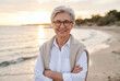 © Meow Creations - Smiling senior woman with short silver hair and glasses standing on a sandy beach during sunset