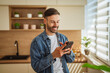 © Miljan Živković - Young man smiling using smart phone in modern kitchen
