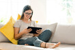 © Westend61 - Smiling brunette woman with glasses relaxing on couch and looking at her tablet