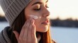 © Pete - Young woman applies face cream outdoors in winter. She wears a grey beanie and scarf, protecting her skin from cold. Healthy glow from moisturizing.
