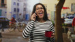 © Krakenimages.com - Middle aged hispanic woman in striped shirt holds phone to ear with one hand and red enamel mug in the other while standing on a busy urban street; concerned.