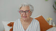 © Krakenimages.com - Elderly woman wearing colorful glasses sits on bed and looks at camera among orange pillows and a vase on a bedside table in building; calm reflection.