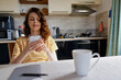 © SHOTPRIME STUDIO - Young woman in a striped yellow tee sits at a bright kitchen table, smiling as she looks at her smartphone, with warm natural light and a casual, relaxed mood.