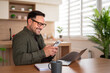 © Moon Safari - Smiling man wearing glasses using a smartphone and laptop, working remotely from a domestic kitchen, embodying modern work life balance and digital connectivity for business