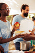 © DC Studio - Close-up shot of generous black woman working in community food bank, serving free meals for the needy. Volunteering team of charity workers helping and sharing nutritrious items to homeless people.