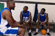 © wavebreak3 - African American male teammates sitting on locker room bench in blue with ball and headphones