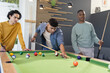 © wavebreak3 - Diverse male friends leaning over pool table lining up shot in modern rec room with cues