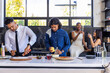 © wavebreak3 - Diverse friends making pizza on kitchen island, man in tie sprinkling sauce, woman using smartphone