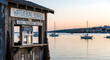 © LaksmanaDPI - Weathered wooden waterfront rentals shack with service window overlooking a calm bay with moored sailboats at sunset