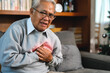 © KL 1981 - Elderly man holds his chest tightly. He shows clear pain and discomfort. His expression is distressed and worried. He sits on a gray sofa. Background includes bookshelves in living room at home